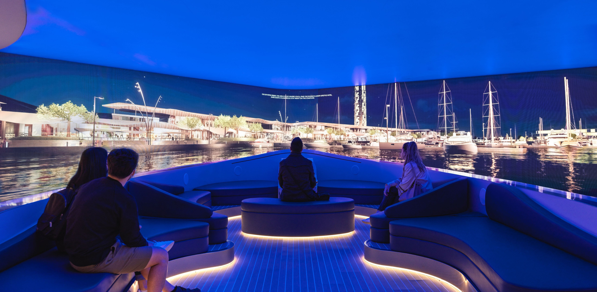 A group of visitors are seated on curved, illuminated benches inside an immersive experience room designed to look like the deck of a yacht. They are watching a panoramic, wrap-around screen displaying a vibrant, photorealistic marina at night.