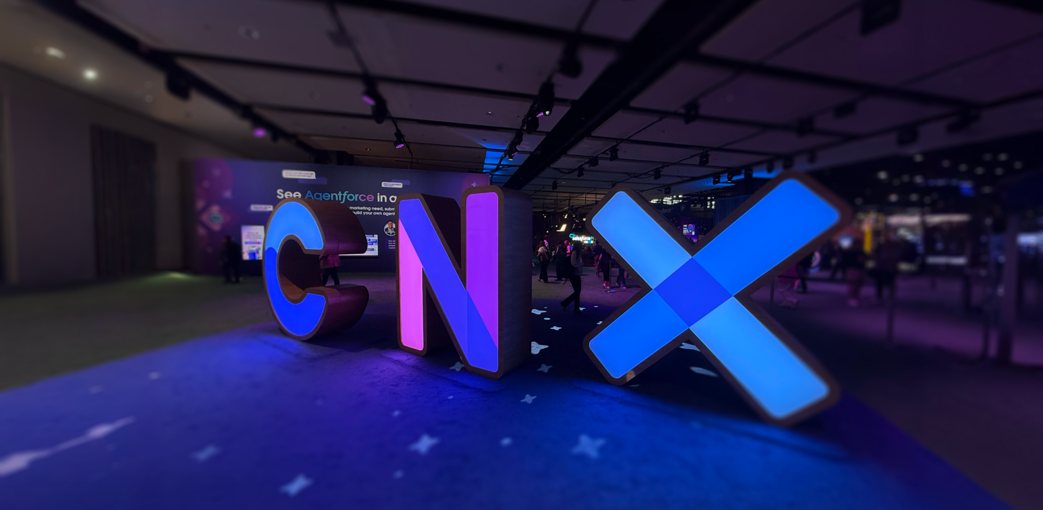 A low-angle, wide shot of large, illuminated, three-dimensional letters spelling "CNX" in a dimly lit event hall. The 'C' glows blue, the 'N' glows purple, and the 'X' glows with a bright blue light. The letters are placed on a dark blue carpet with a subtle pattern, and they cast colorful reflections on the floor. In the blurred background, people can be seen walking around the conference space.