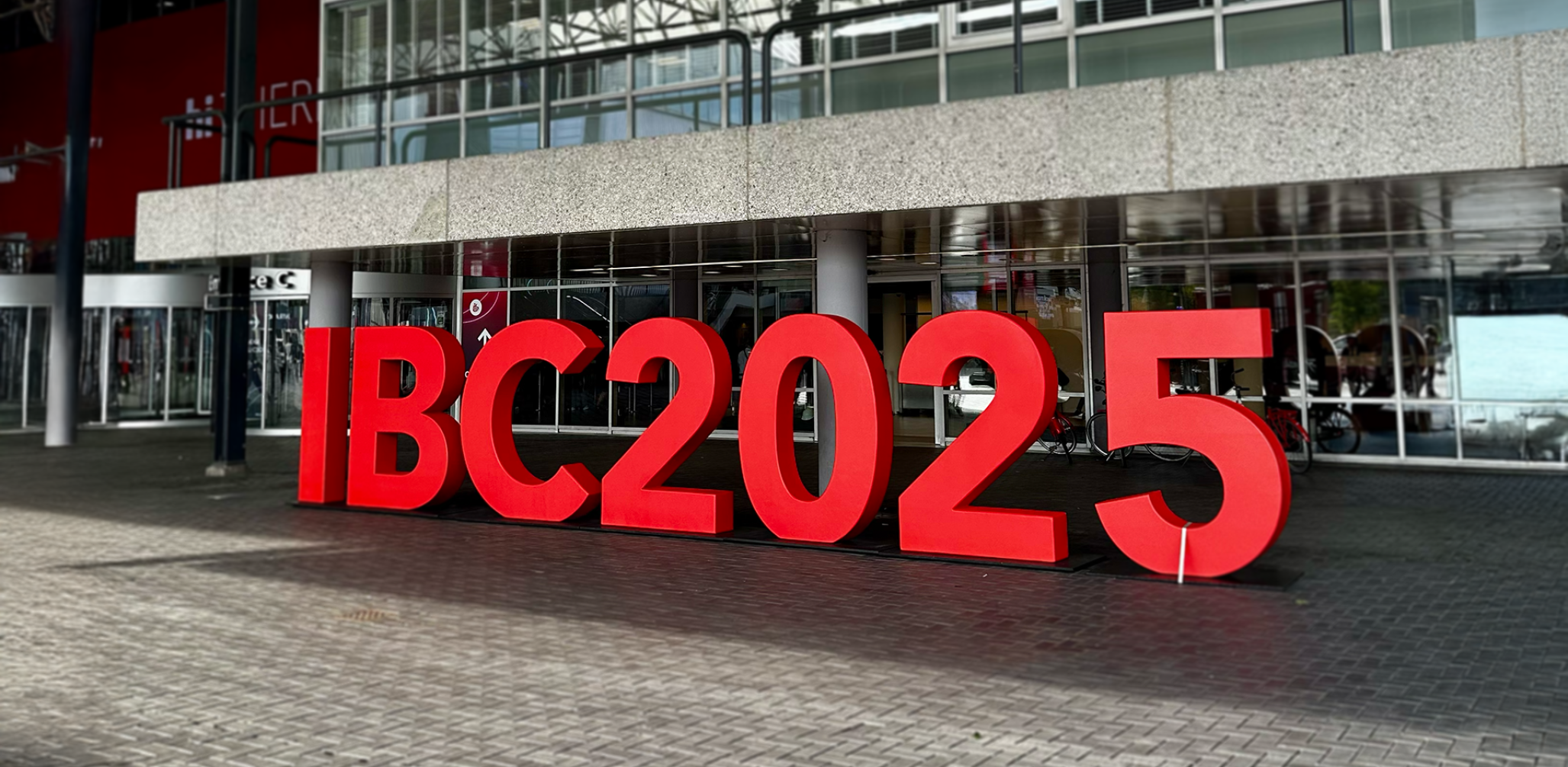 Large, three-dimensional red letters spelling "IBC2025" stand on a brick plaza in front of the entrance to a modern convention center with a glass facade.