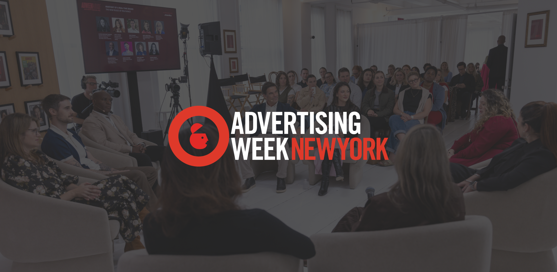 A panel discussion in progress at an event, with the "Advertising Week New York" logo overlaid in the center. In the background, a group of panelists sits in a semi-circle of chairs facing an audience, with a large screen displaying speaker headshots on the wall.