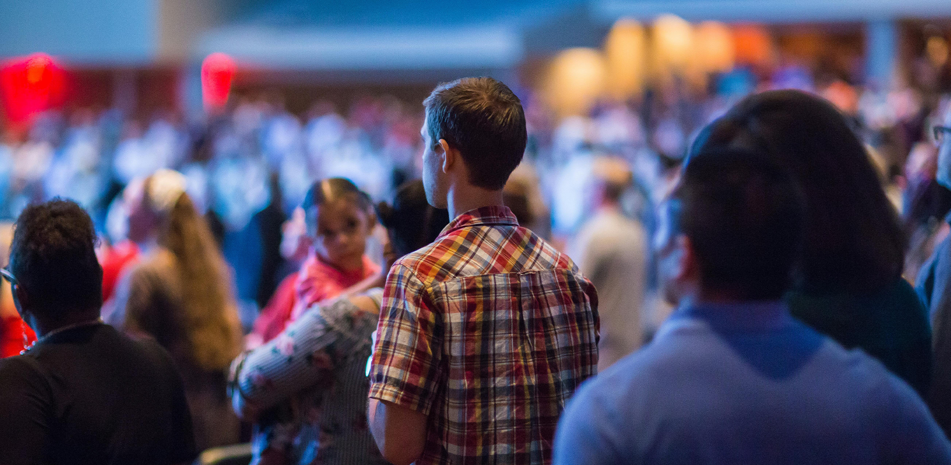 A rear view of a standing crowd in a large, dimly lit indoor venue, focusing on a man in a plaid shirt in the center. To his left, a woman holds a small child who is looking sideways, while the foreground and background contain other attendees blurred out of focus.