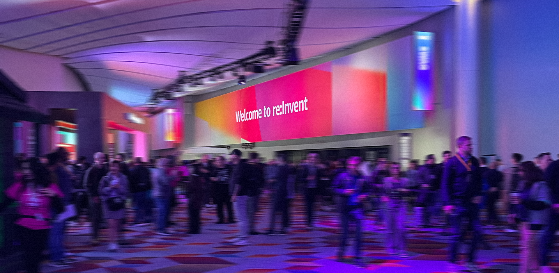 A photograph of a large, crowded convention center hall. A large, curved sign with a colorful pink, orange, and purple gradient background reads "Welcome to re:Invent". The space is illuminated with purple and blue lights, and the floor has a geometric pattern. Numerous attendees are walking around the hall.