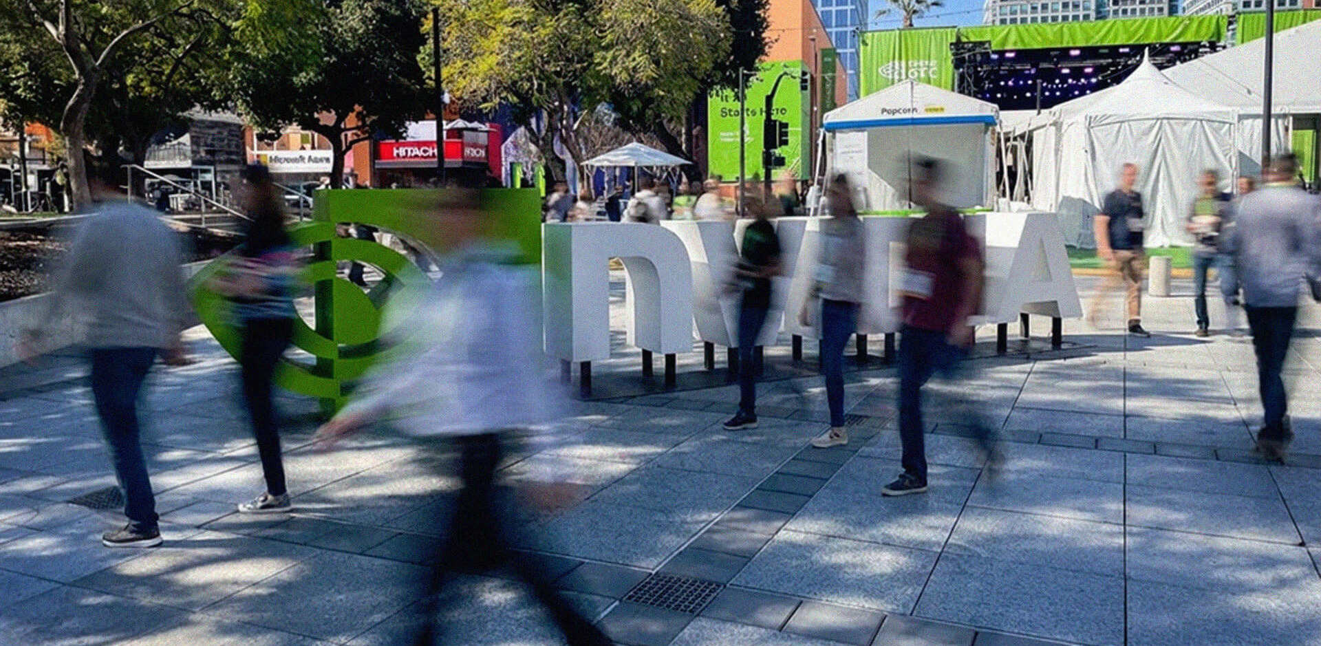 A wide-angle, slightly blurred shot of an outdoor plaza at the NVIDIA GTC 2026 conference in San Jose. Large, 3D white letters spelling out "NVIDIA" stand in the center, with the green NVIDIA logo to the left. People are captured in motion, appearing as blurred figures walking across the stone-tiled ground, creating a sense of a busy, electric atmosphere. In the background, there are green banners, white event tents, trees, and city buildings under a clear blue sky.