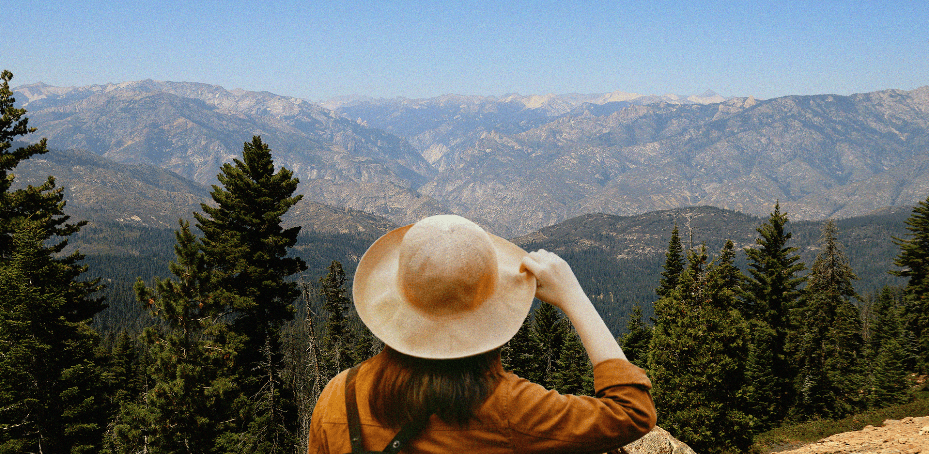A person seen from behind wearing a wide-brimmed tan hat and an ochre jacket, gazing out at a vast, rugged mountain range and evergreen forest under a clear blue sky.