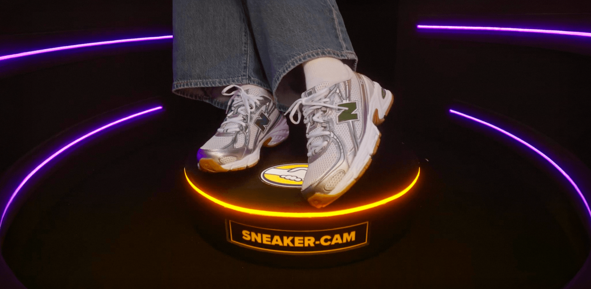 A close-up of a person's feet wearing blue jeans, white socks, and silver and white New Balance sneakers featuring a green "N" logo. They are standing on a small, circular black platform that has a glowing orange ring light around its top edge. The front of the platform displays a yellow label reading "SNEAKER-CAM".