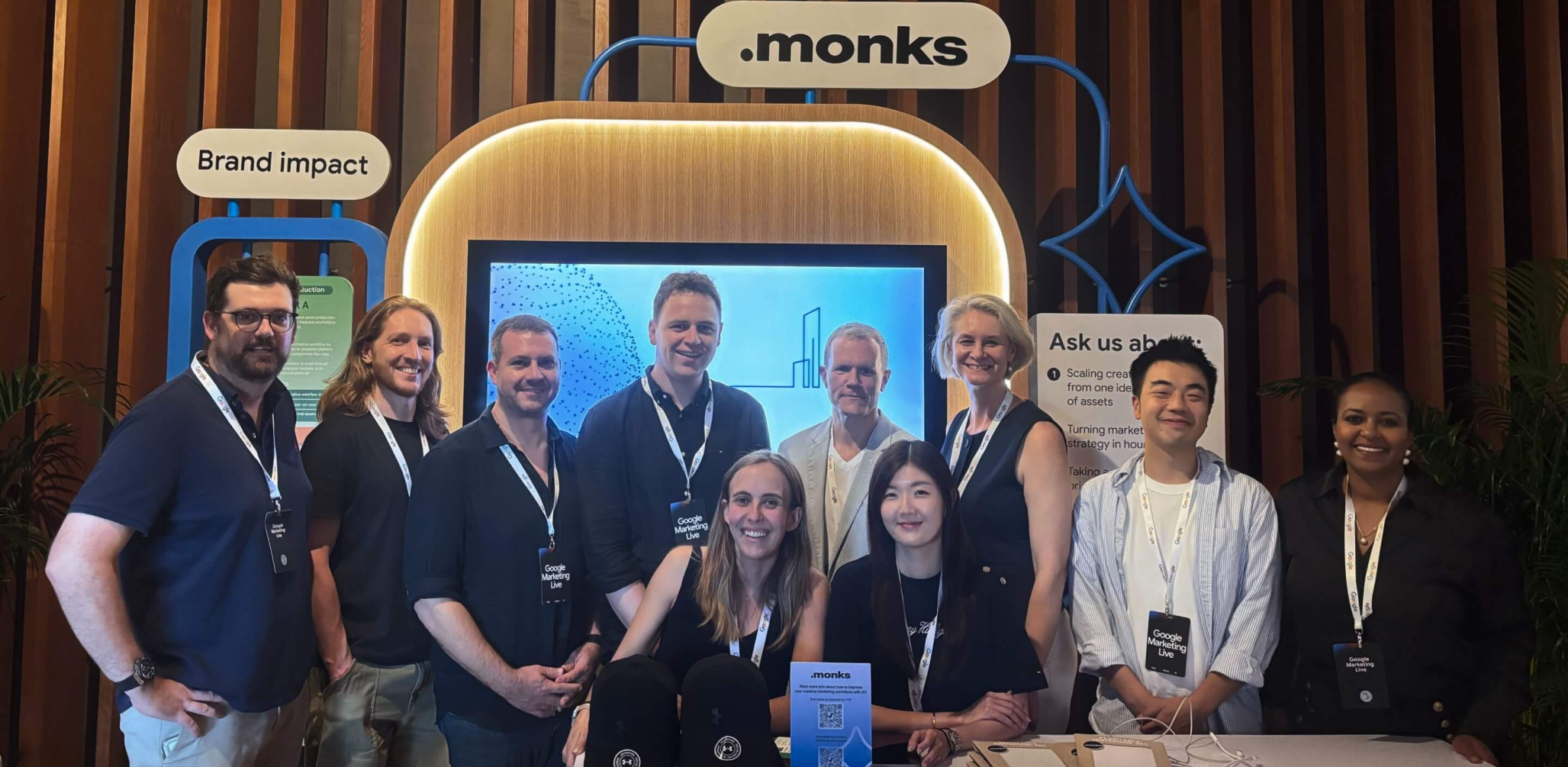 A group of ten smiling professionals posing together at a ".monks" event booth. The attendees are wearing lanyards with "Google Marketing Live" badges. Behind them is a large display screen, an illuminated ".monks" logo, and booth signs reading "Brand impact" and "Ask us about:". In the foreground, a table is set up with branded black caps, brochures, and a small sign featuring a QR code.