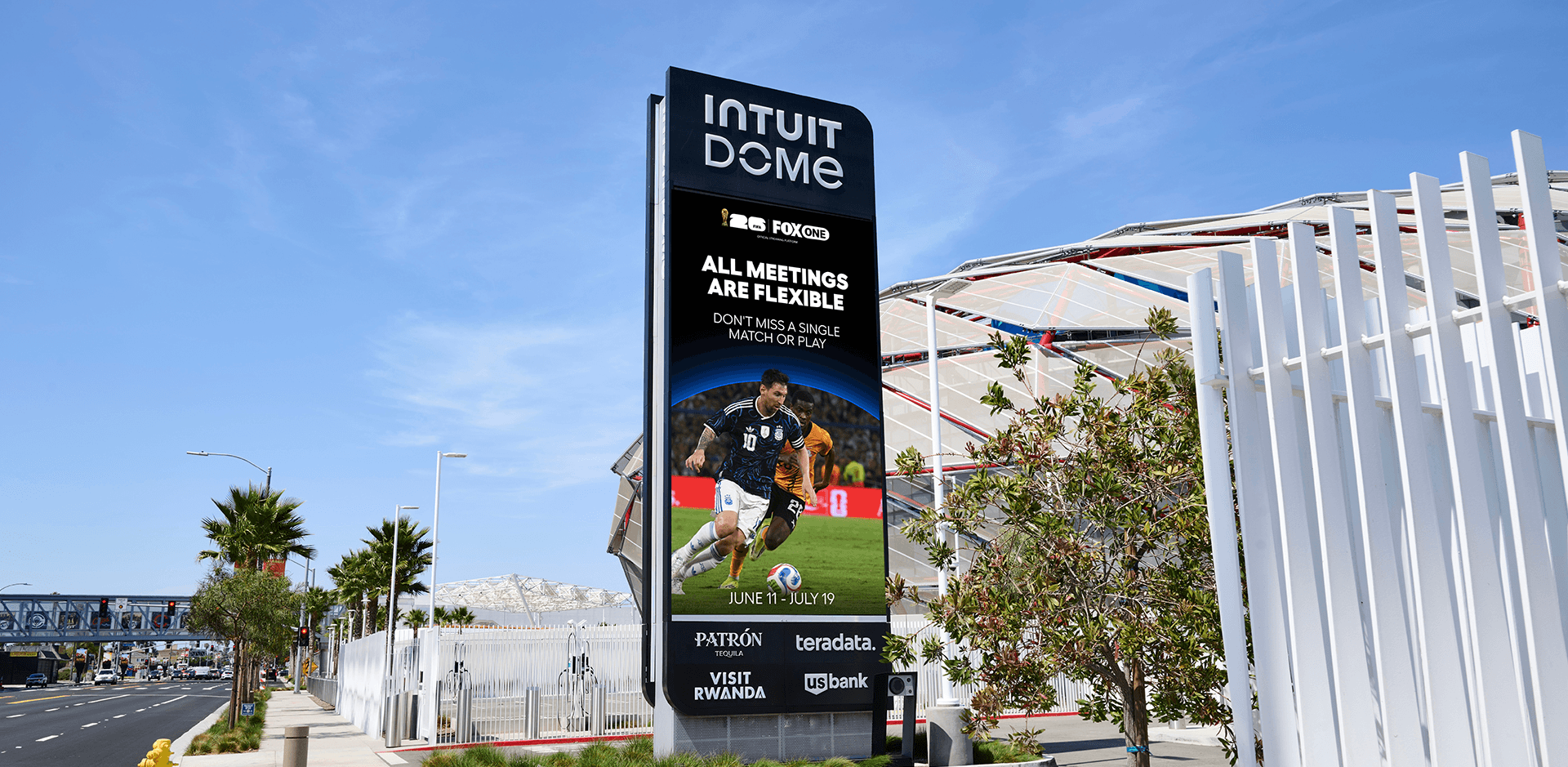 A tall digital billboard outside the Intuit Dome displaying a FIFA World Cup advertisement featuring soccer player Lionel Messi in action. The screen reads "ALL MEETINGS ARE FLEXIBLE" above the dates June 11 through July 19, alongside logos for Fox and various corporate sponsors like Patron and US Bank. The sign stands on a sunny, palm-tree-lined sidewalk with the stadium's exterior visible in the background behind a white fence.