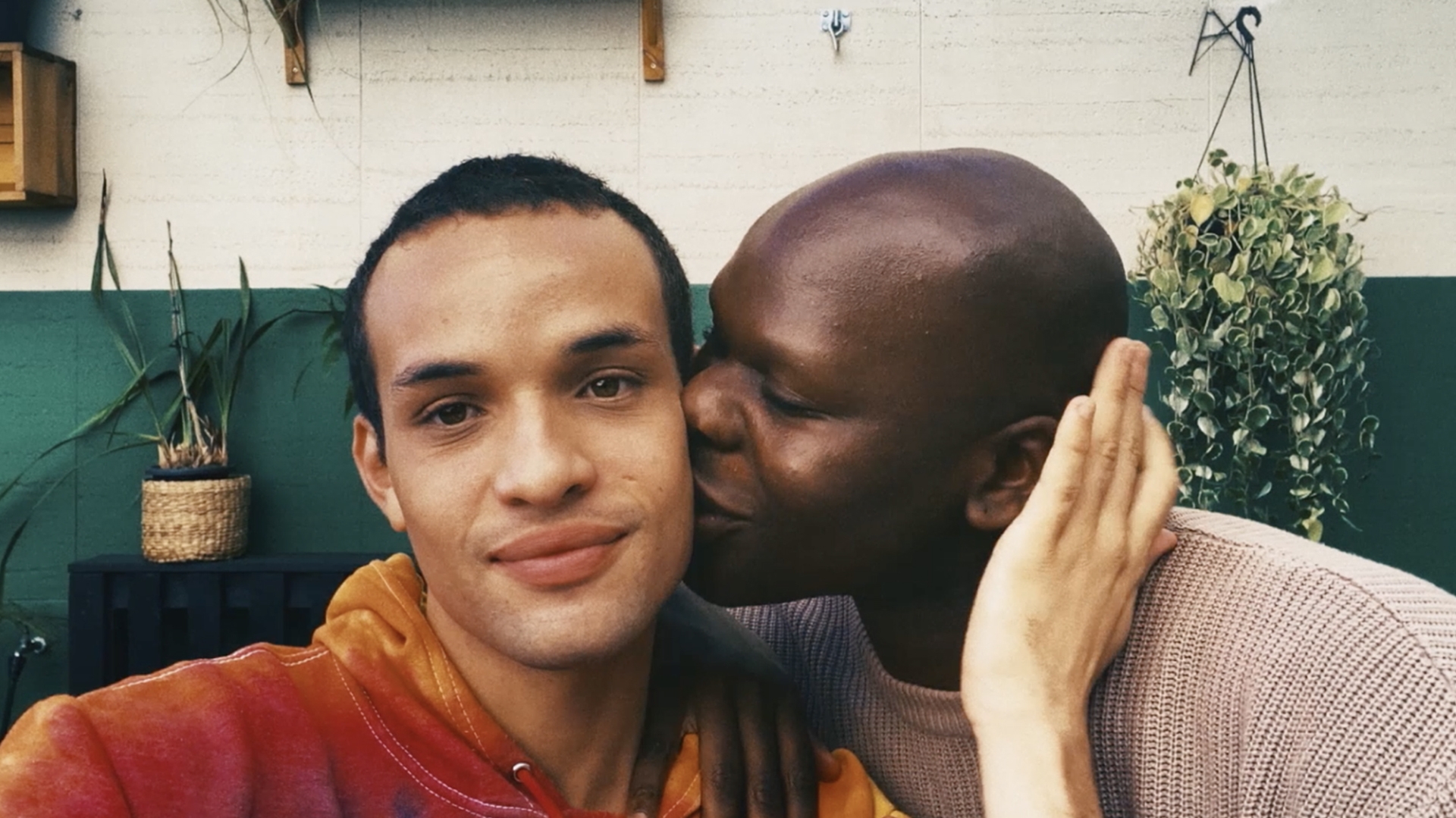 two men next to each other, one facing the camera while the other kisses the first on the cheek