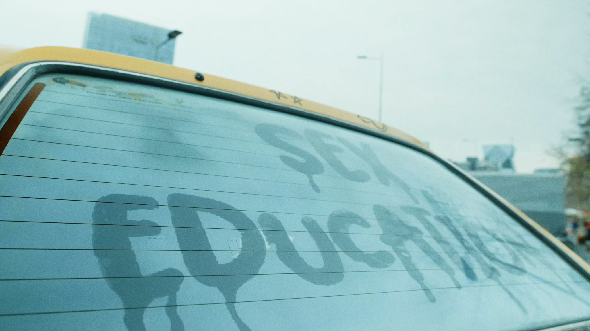 The back window of a car is all condensed from the inside. Somebody wrote 'Sex Education' on the condensation on the window.