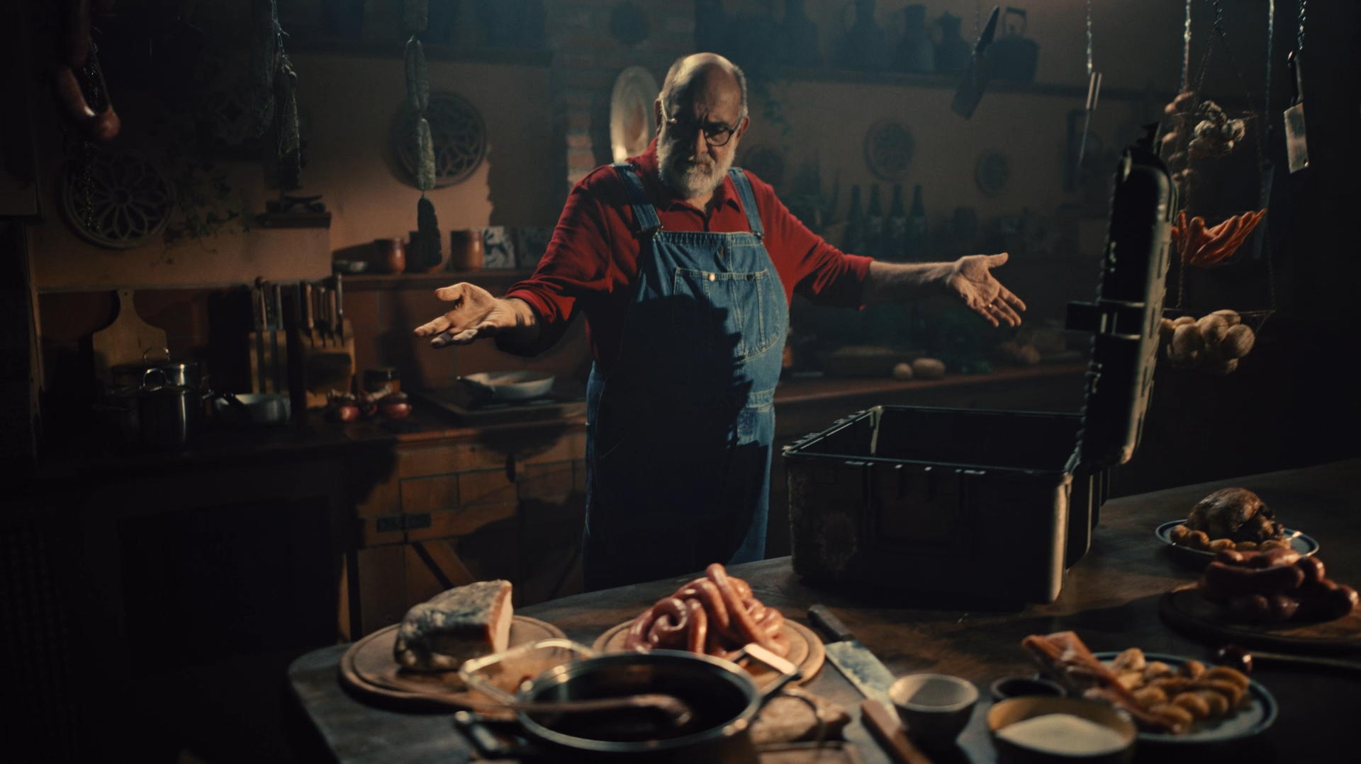 Dark setting with a opened chest on the table. A man in dungarees stading behind a tabl spreading his hands wide open