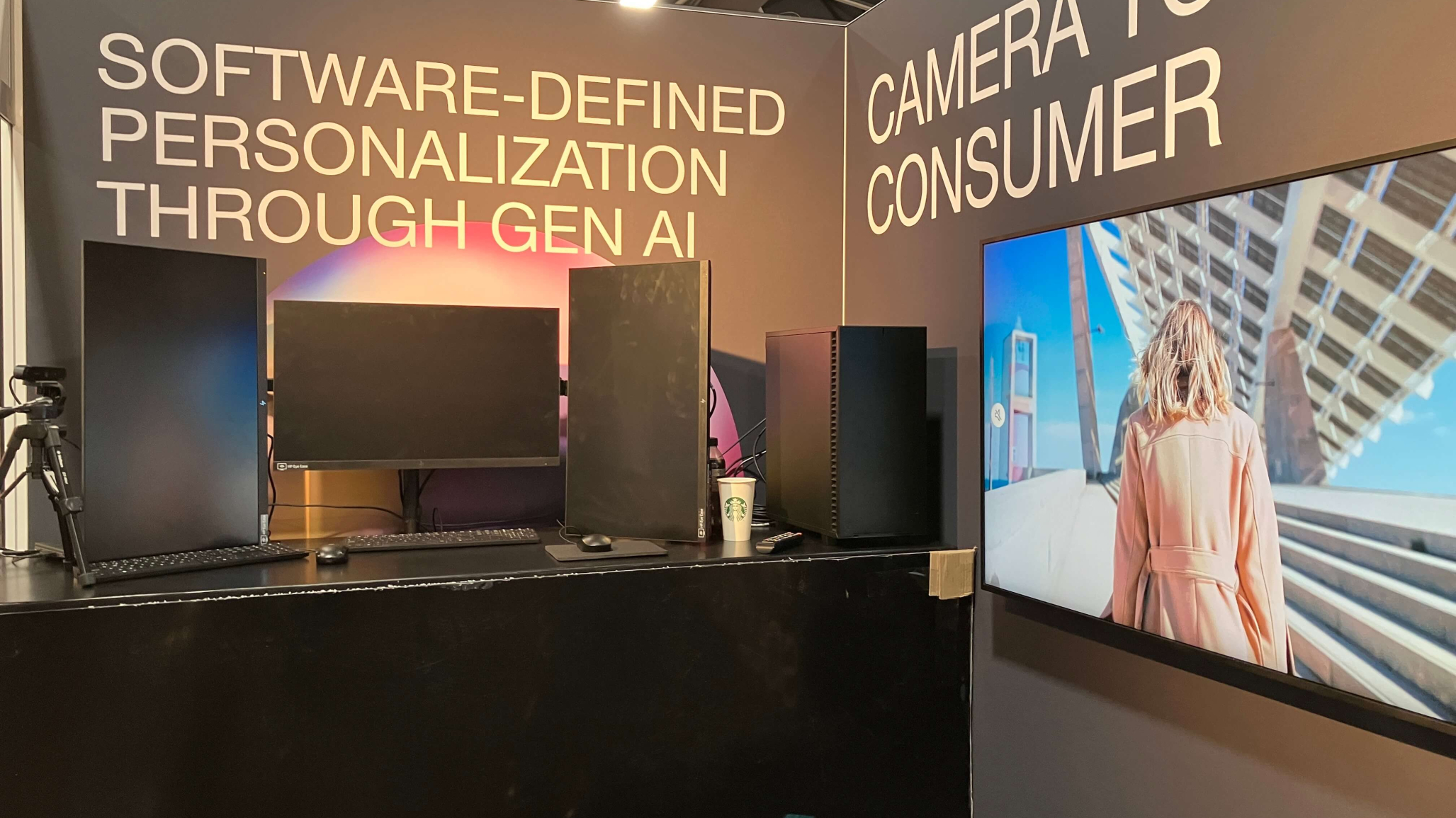 The Monks booth at IBC. The booth displays the text "Software-Defined Personalization Through Gen AI" and "Camera to Consumer." The setup includes three computer monitors. To the right, a large screen shows an image of a person wearing a coat, standing outdoors near a modern architectural structure.