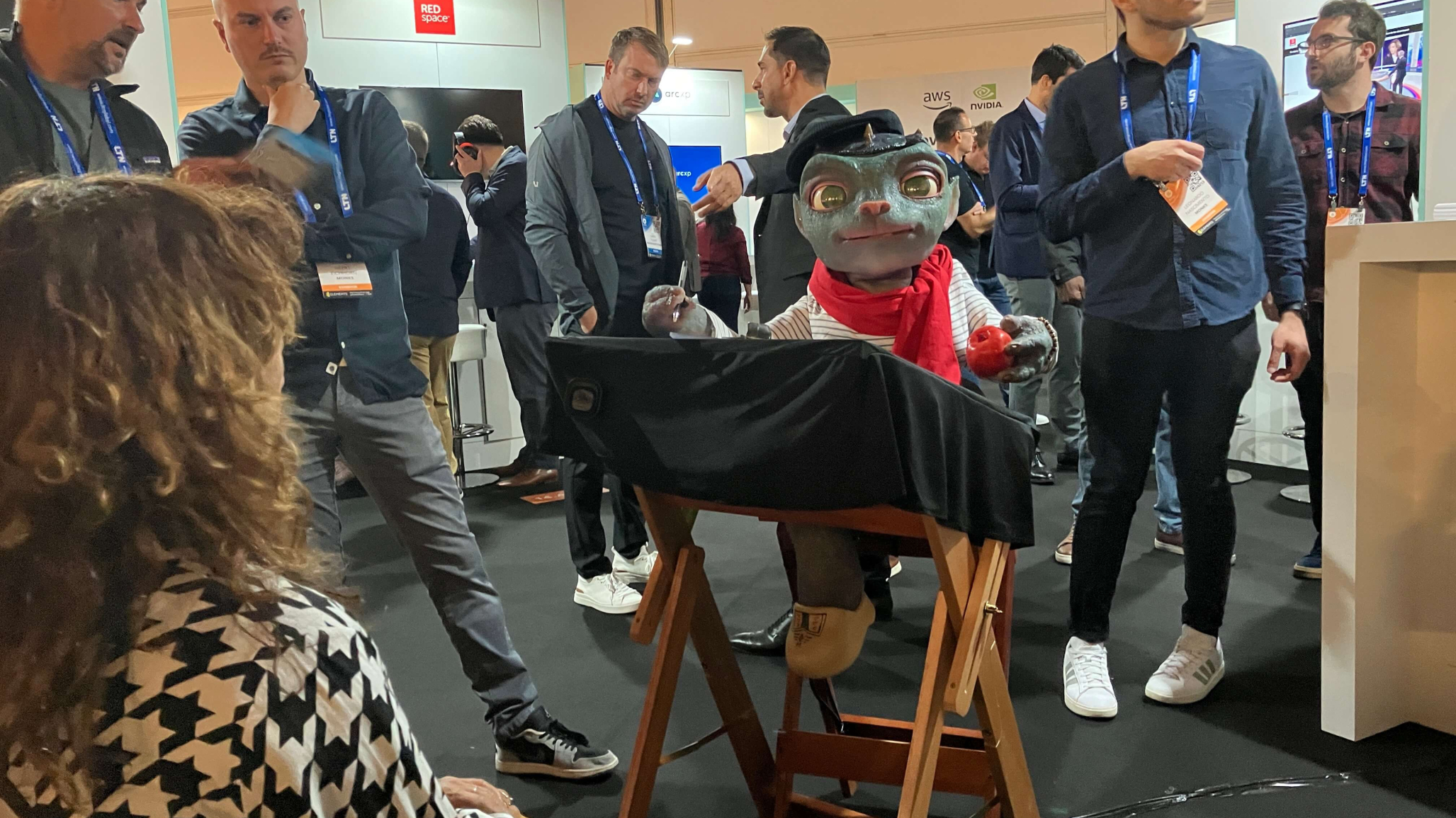 A group of people gather around a robotic alien dressed in a beret and red scarf, holding a red apple, at a trade show. The puppet is seated at a small table, engaging with a seated woman in the foreground by drawing her portrait. Various exhibitors and displays are visible in the background.