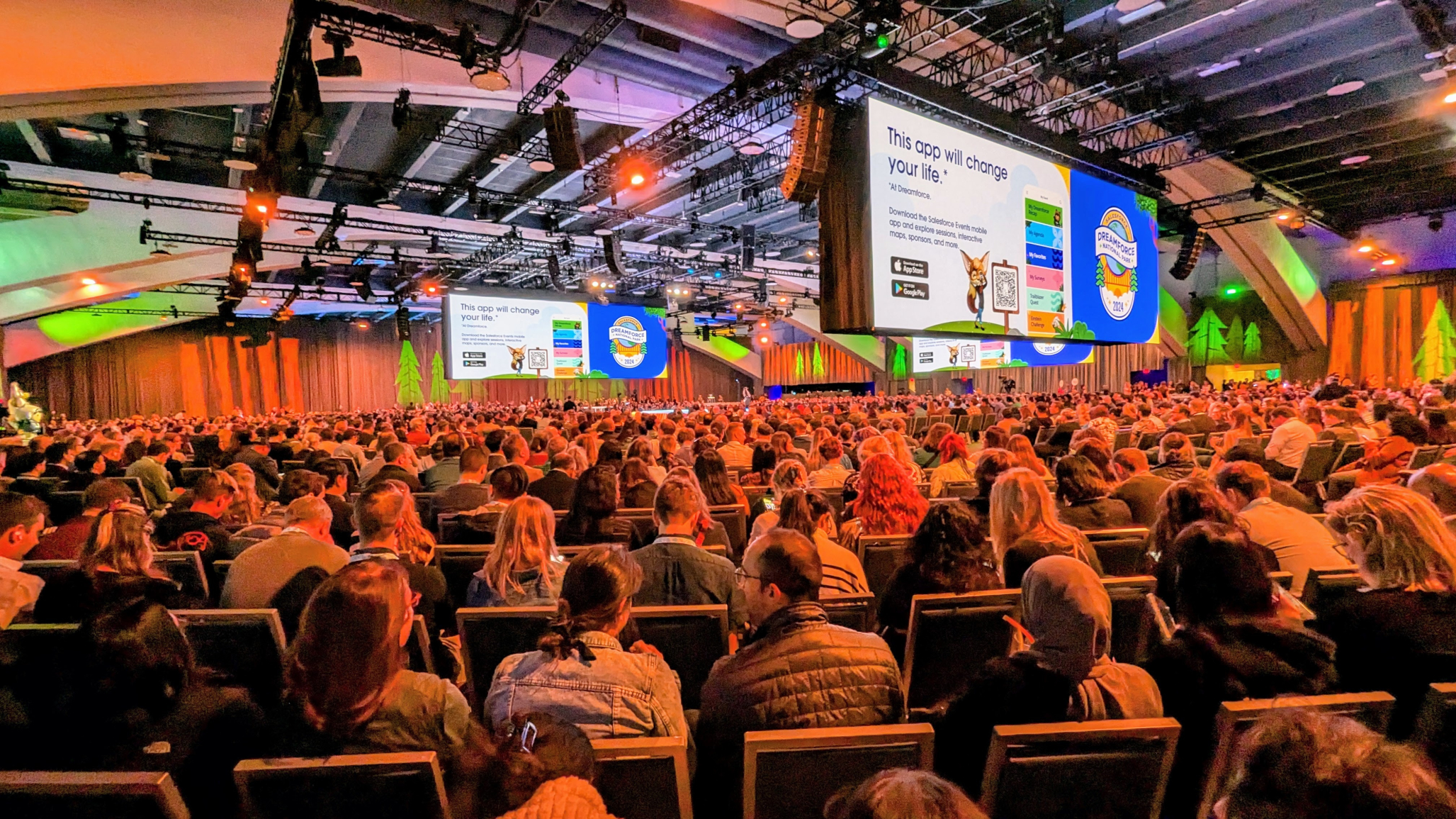 A large conference room filled with attendees at Dreamforce. People are seated in rows, facing a stage with two large screens displaying presentations. The room is illuminated with colorful lighting, and banners with conference branding are visible. The atmosphere is lively and focused.