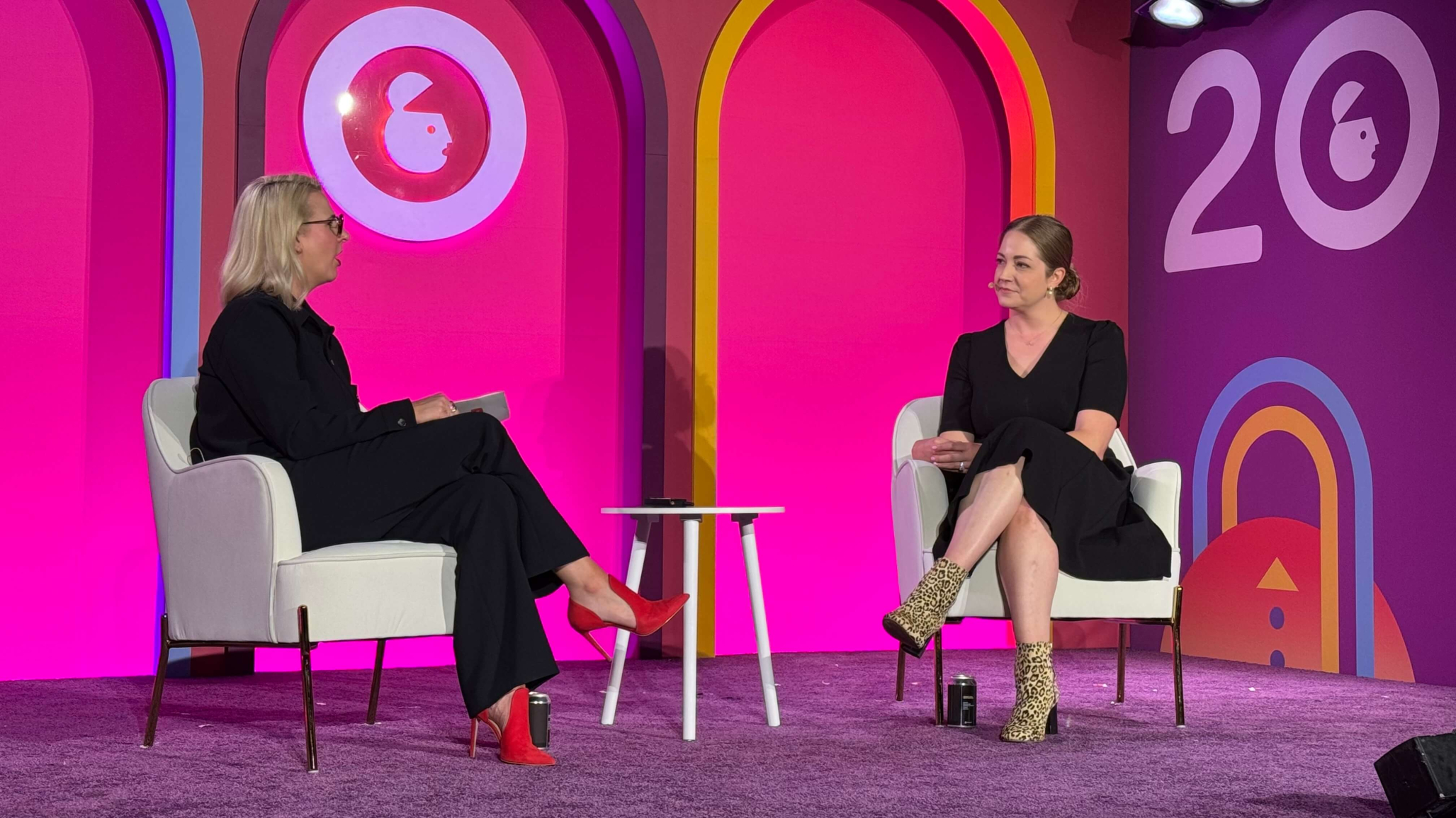 Two women are seated on a stage with a vibrant pink and purple backdrop featuring the number "20" and stylized graphics. Noël Paasch sits on the left; Brittany Blanchard, on the right.