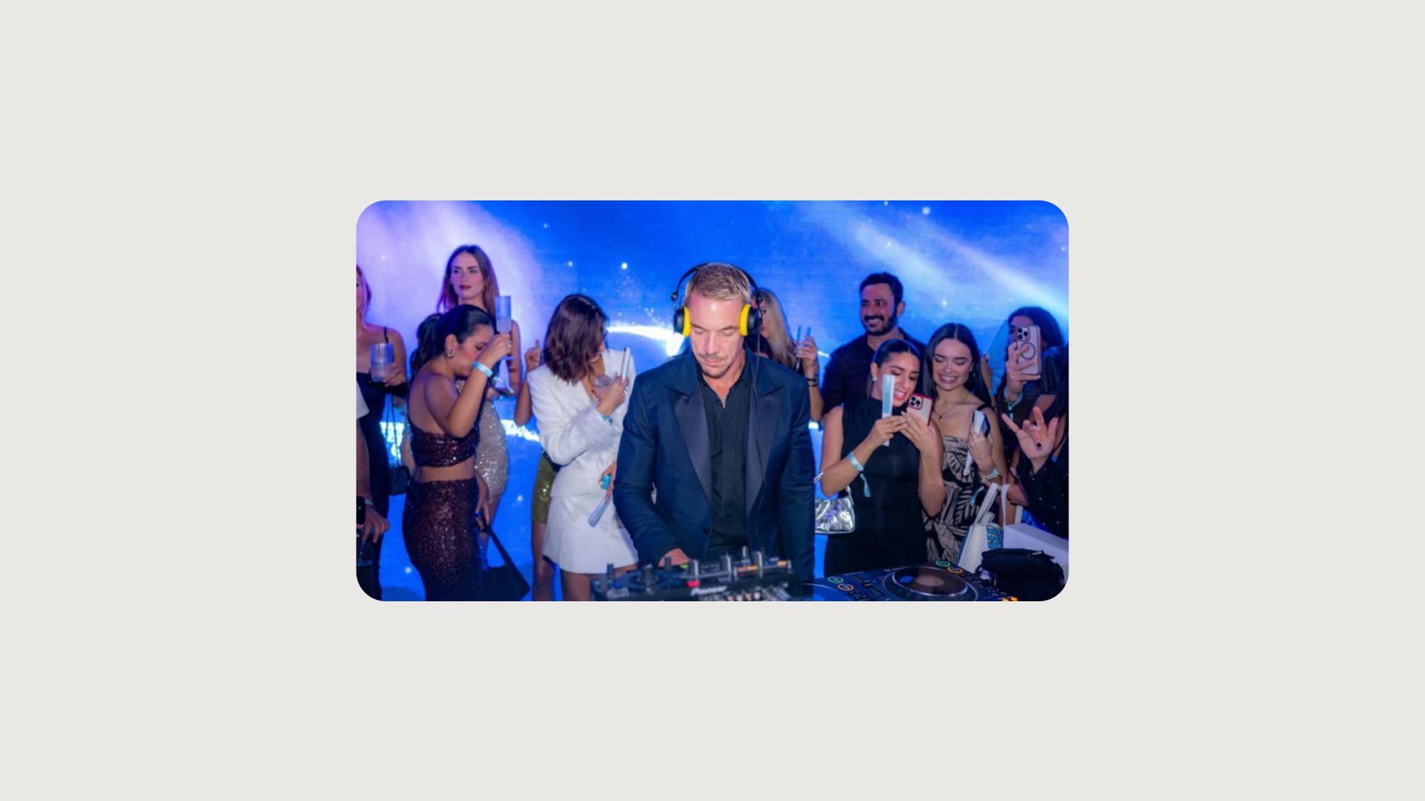 A DJ in a black suit and headphones smiles while performing on a brightly lit blue stage, surrounded by a crowd of people dancing and taking photos with their phones.