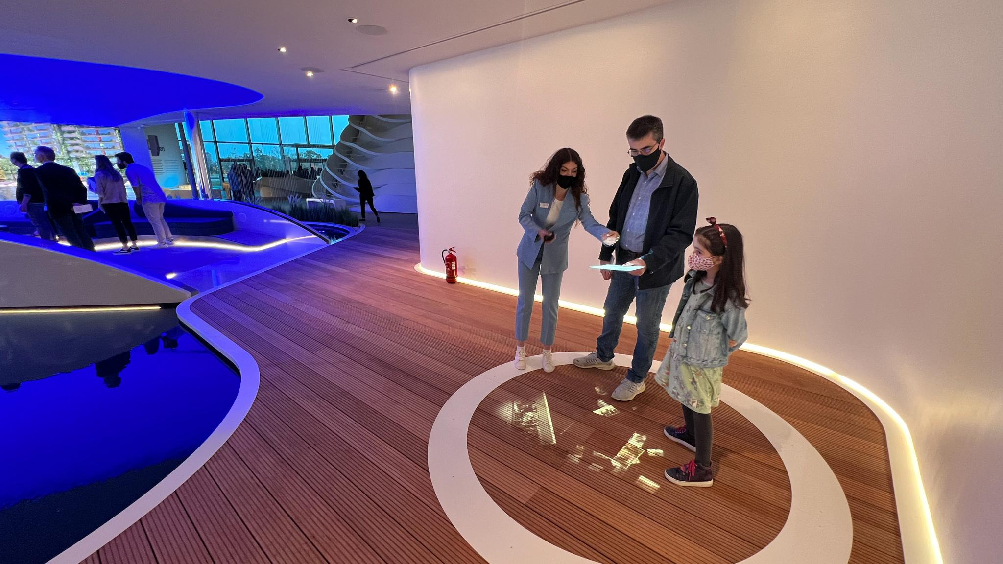 A family of three, a man, woman, and young girl, all wearing face masks, stand in a modern-looking room with wooden floors and curved, illuminated white walls. They are gathered around a tablet that the woman is holding, standing within a large white circle embedded in the floor.