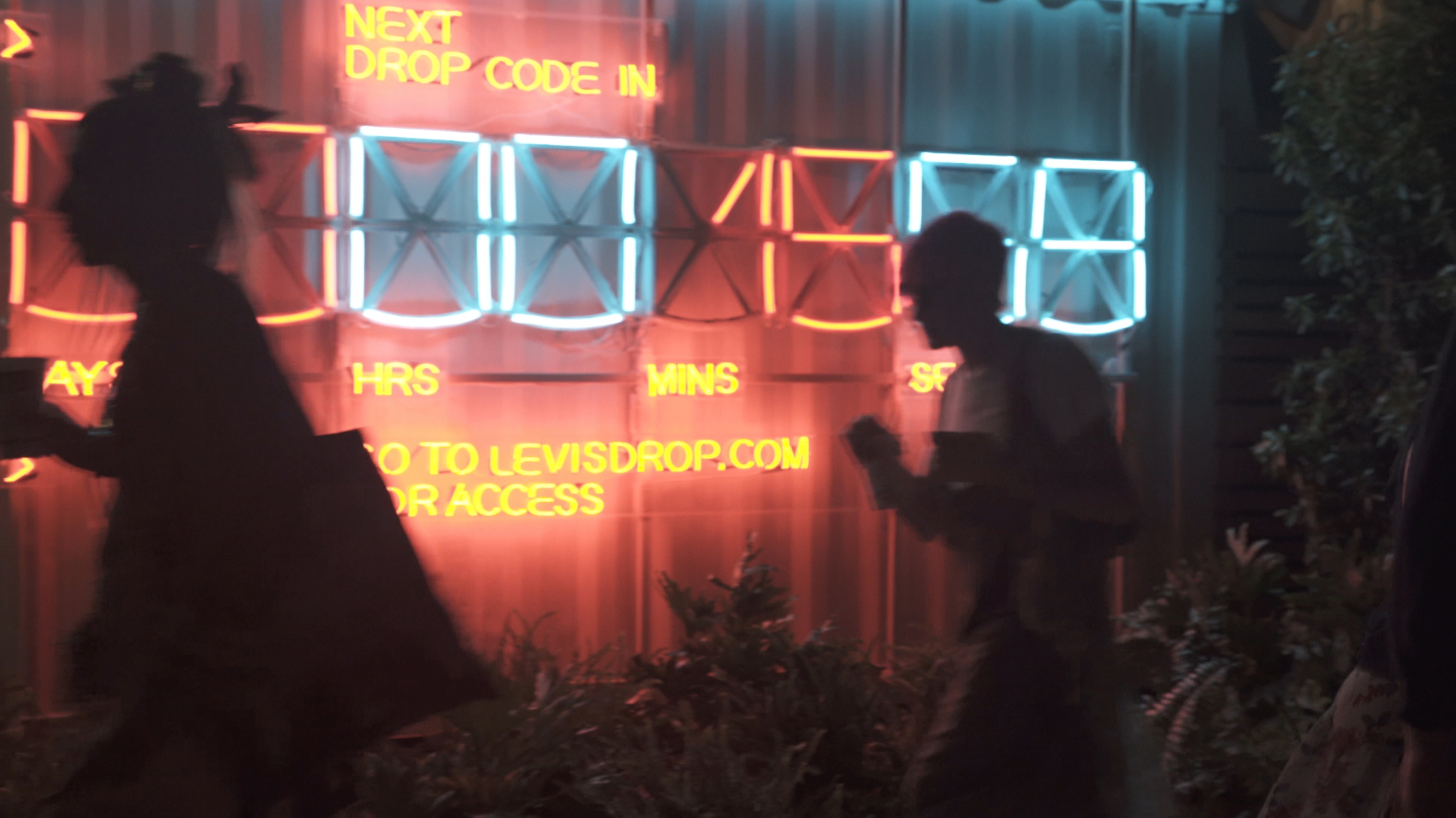 A nighttime scene of a large, digital countdown clock with glowing orange and blue neon numbers, set up for a Levi's apparel drop. In the foreground, silhouettes of people walk past the sign.