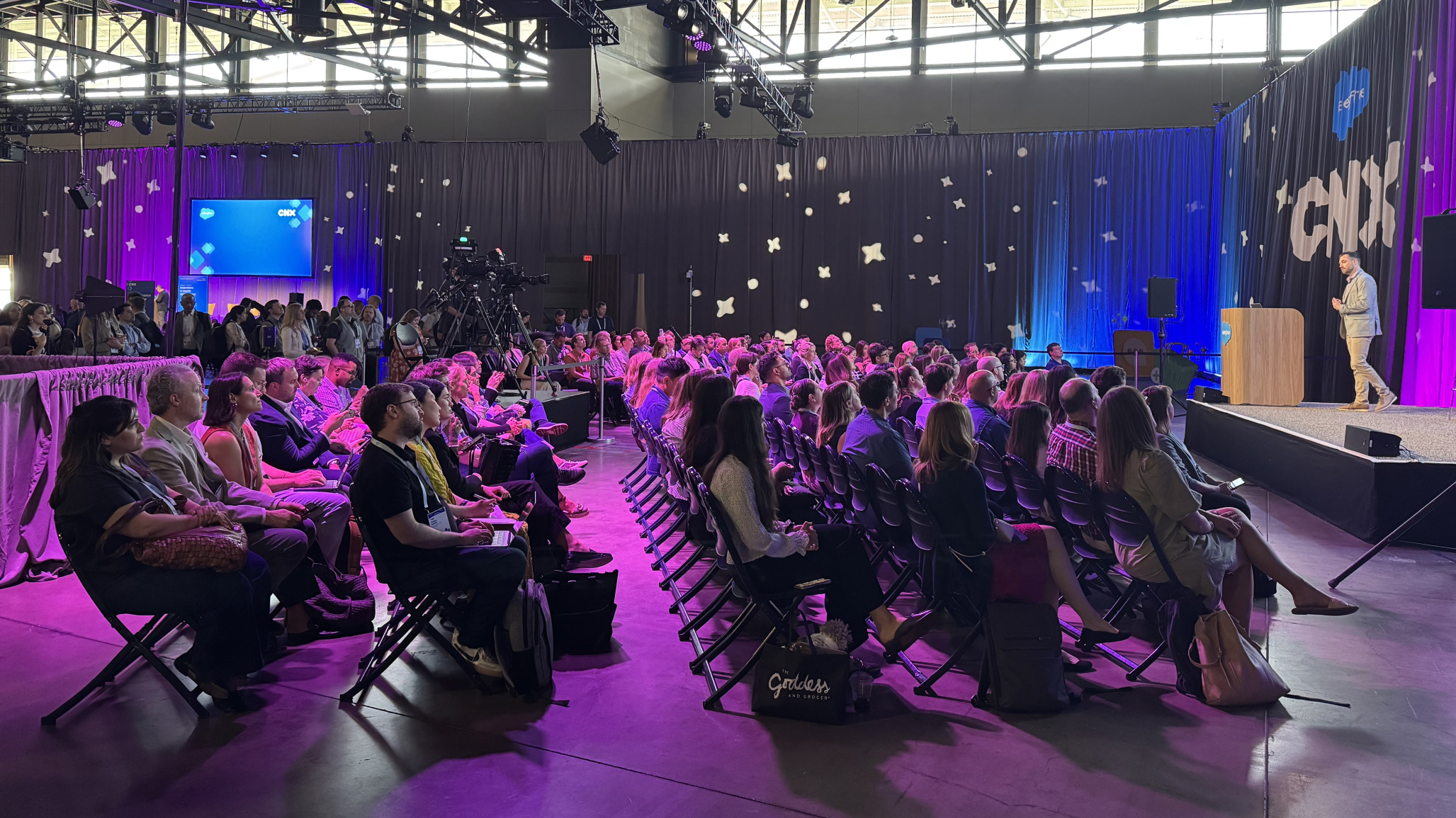 A wide shot of a conference hall where a man is speaking on a stage to a large, seated audience. The room is dimly lit with dramatic purple and blue stage lights. The backdrop behind the speaker is a dark curtain with white star-like cutouts and the large letters "CNX" on the right. The audience is attentively watching the speaker from rows of chairs.