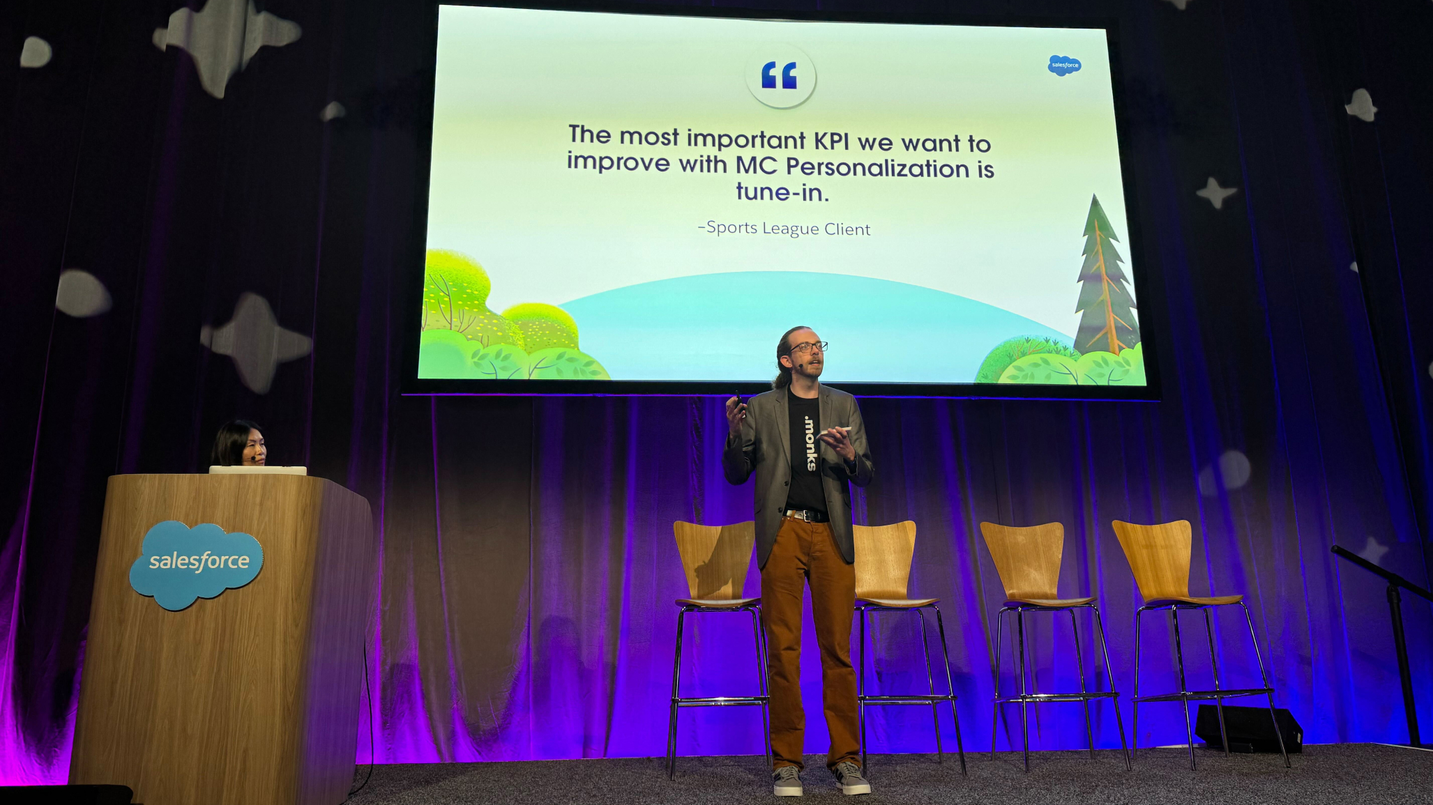 Nathan is on a conference stage, holding a microphone and presenting. To his left, a woman peeks over a wooden lectern that has a Salesforce logo on it. Behind them, a large screen displays a presentation slide with a quote that reads: "'The most important KPI we want to improve with MC Personalization is tune-in.' - Sports League Client." The stage is lit with blue and purple lights and has three empty wooden chairs to the right of the speaker.