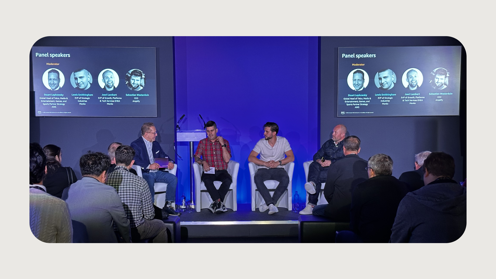 A panel of four male speakers sits on a stage, addressing an audience. The two men on the left are seated on white armchairs, while the two on the right are seated on white stools. Behind them, two large screens display headshots and names of "Panel speakers" with "Moderator" at the top. The stage is lit with blue light, and the audience, mostly men, are visible from behind, facing the stage.