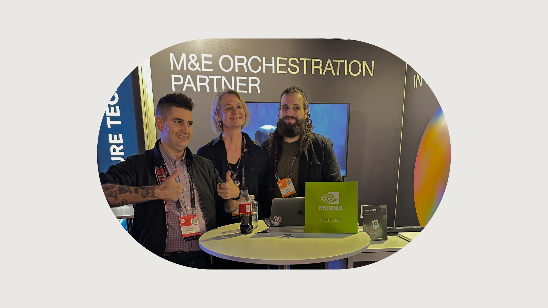 Two men and a woman pose smiling at a conference booth. The man on the left and the woman in the middle are both giving a thumbs-up. They stand behind a table that features a green sign with the NVIDIA Partner logo. In the background, a large sign reads, "M&E ORCHESTRATION PARTNER."
