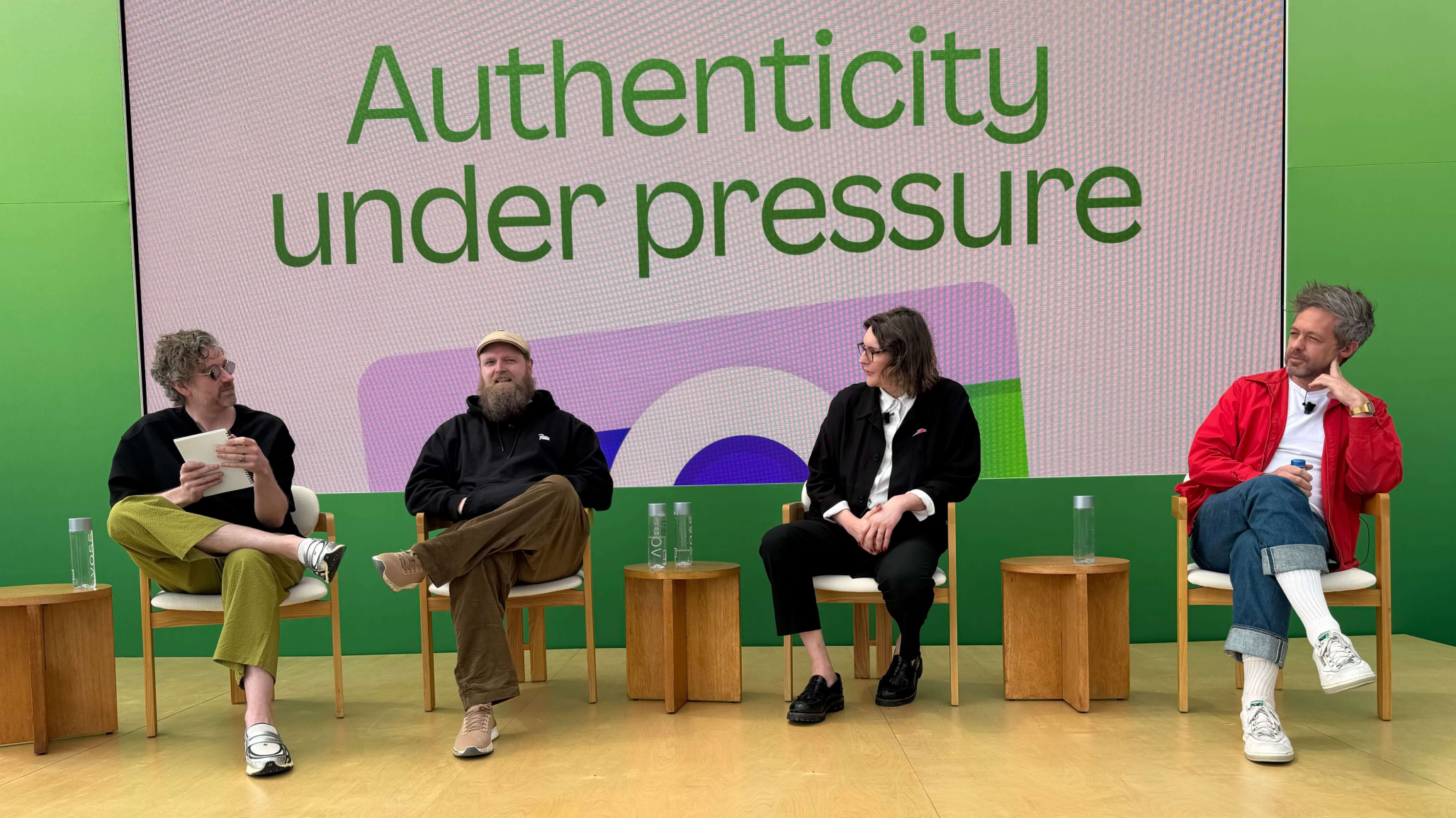 A panel of four people seated on wooden chairs on a stage for a discussion titled "Authenticity under pressure," which is displayed in large green text on a screen behind them. The participants, three men and one woman, are dressed in casual, modern attire. Small wooden side tables with water bottles are positioned between the speakers against a vibrant green backdrop.