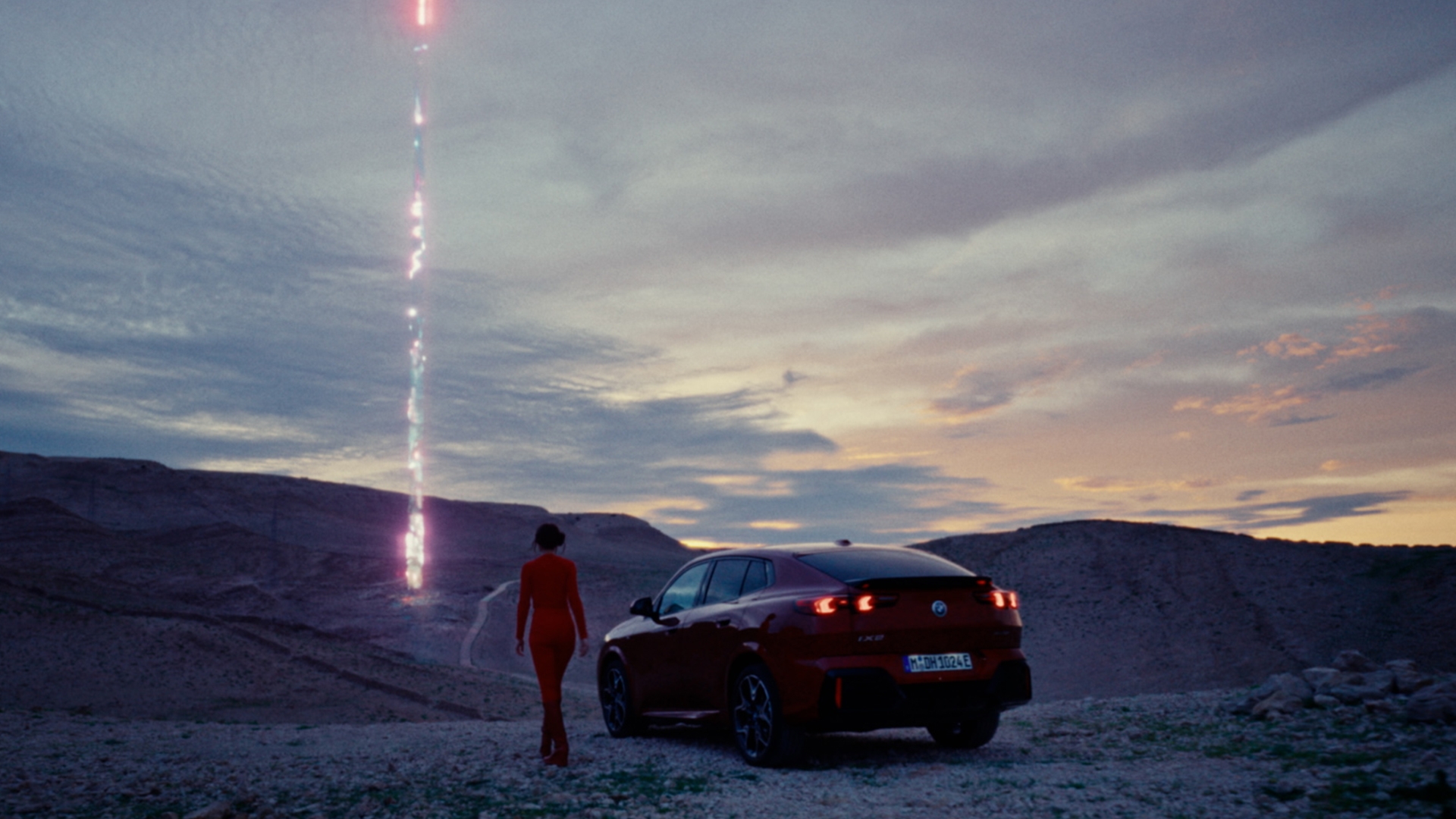 Lil Miquela stands next to the new BMW X2 and the car is in the desert. In the distance you see a futuristic beam of light coming from the sky to the ground.