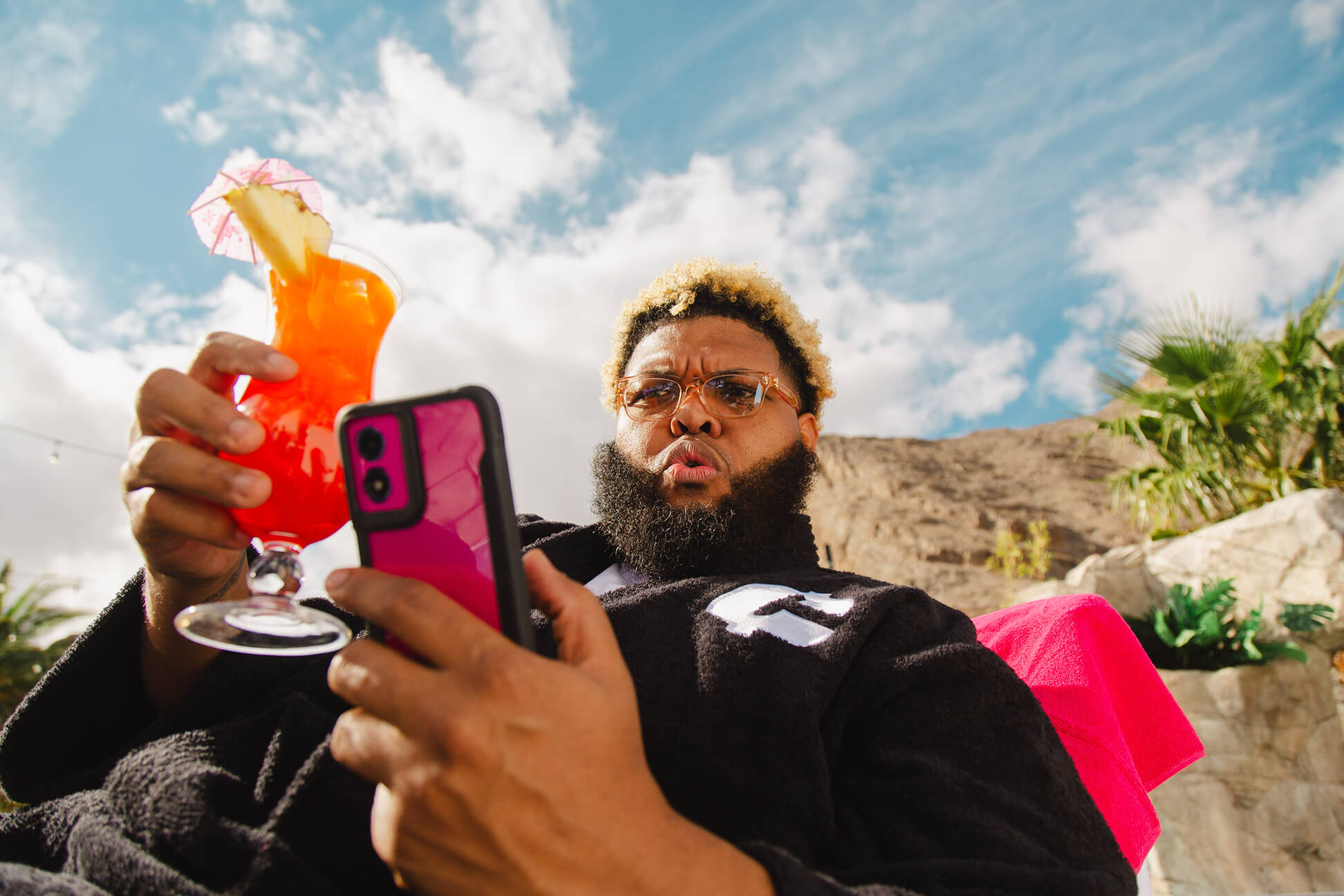 Influencer Druski sitting in a lounge chair at a pool with a T-Mobile phone in one hand and a cocktail in another.