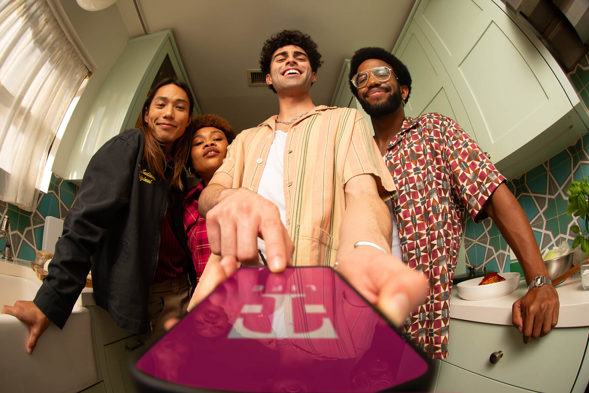 4 T-Mobile users standing together in a kitchen looking at a phone with one of them holding a phone that has the T-Mobile app open on it.