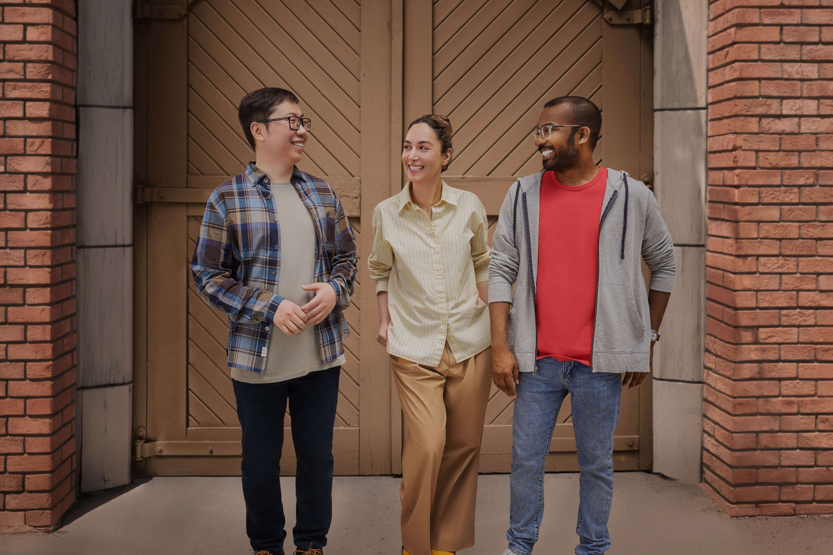 Three casually dressed people—two men and a woman—standing together in front of large wooden double doors framed by brick pillars. They are smiling and looking at each other as if sharing a friendly conversation. From left to right: a man in a plaid shirt and glasses, a woman in a light yellow patterned button-down shirt and tan pants, and a man wearing glasses, a red t-shirt, and a grey zip-up hoodie.