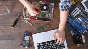 A laptop siting on a table with computer hardware lying around while someone works on it