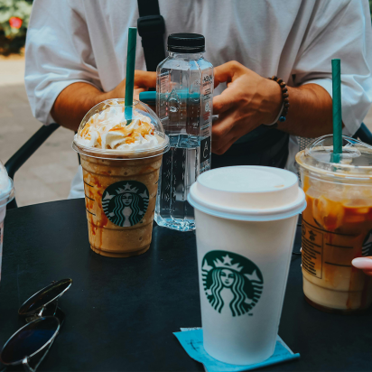 A table outside with all sorts of Starbucks coffe cups on it. At the back of the table you see two hands of a person who is using his mobile.