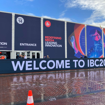 The facade of the IBC convention hall is depicted. Large banners outside display text reading "Badge Collection," "Entrance," and "Redefining Media Leading Innovation." A futuristic image of a person wearing a virtual reality headset is featured. The foreground has a sign that reads "Welcome to IBC2024." People are gathered near the entrance, and a traffic cone is visible on the wet pavement.