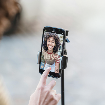 A close-up image of a smartphone mounted on a tripod, capturing a cheerful woman with curly hair wearing a striped shirt during a video recording. A hand is visible in the foreground, about to tap a button on the phone screen.