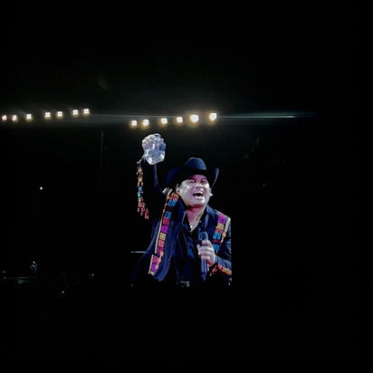 Julión Álvarez, wearing a black cowboy hat and a colorful embroidered jacket, performs on stage at SoFi Stadium. He is smiling, holding a microphone in one hand and raising a bottle in the other. Bright stage lights shine behind him, and the background is mostly dark.