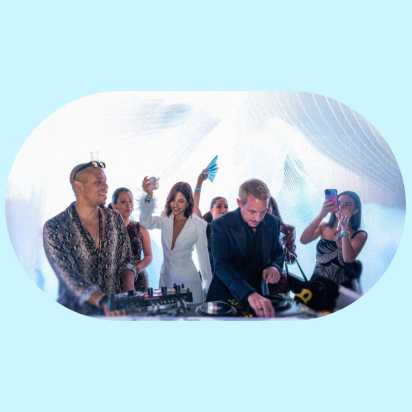 A DJ in a dark suit plays music at a brightly lit event, while another individual in a patterned shirt and sunglasses stands to his left at the DJ booth. Several people are visible behind them, dancing and holding up their phones.