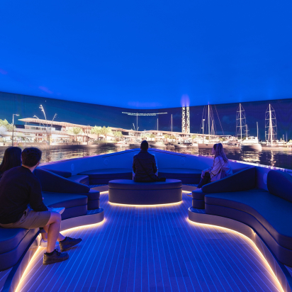 A group of visitors are seated on curved, illuminated benches inside an immersive experience room designed to look like the deck of a yacht. They are watching a panoramic, wrap-around screen displaying a vibrant, photorealistic marina at night.