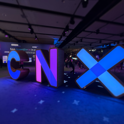 A low-angle, wide shot of large, illuminated, three-dimensional letters spelling "CNX" in a dimly lit event hall. The 'C' glows blue, the 'N' glows purple, and the 'X' glows with a bright blue light. The letters are placed on a dark blue carpet with a subtle pattern, and they cast colorful reflections on the floor. In the blurred background, people can be seen walking around the conference space.