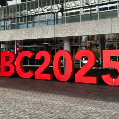 Large, three-dimensional red letters spelling "IBC2025" stand on a brick plaza in front of the entrance to a modern convention center with a glass facade.