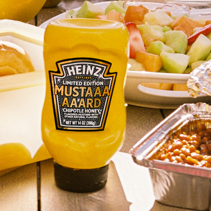 Bright yellow Heinz Limited Edition Mustaaaaard Chipotle Honey bottle, prominently displayed on a wooden picnic table next to a fruit salad, baked beans, and fresh buns, highlighting the perfect summer BBQ condiment.