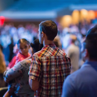 A rear view of a standing crowd in a large, dimly lit indoor venue, focusing on a man in a plaid shirt in the center. To his left, a woman holds a small child who is looking sideways, while the foreground and background contain other attendees blurred out of focus.