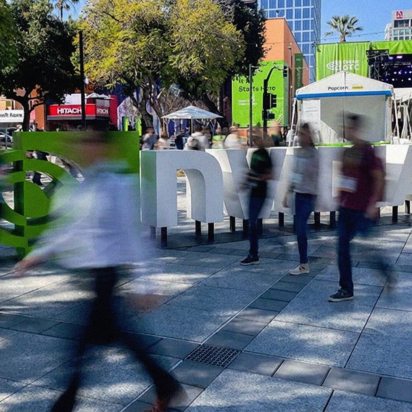 A wide-angle, slightly blurred shot of an outdoor plaza at the NVIDIA GTC 2026 conference in San Jose. Large, 3D white letters spelling out "NVIDIA" stand in the center, with the green NVIDIA logo to the left. People are captured in motion, appearing as blurred figures walking across the stone-tiled ground, creating a sense of a busy, electric atmosphere. In the background, there are green banners, white event tents, trees, and city buildings under a clear blue sky.