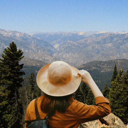 A person seen from behind wearing a wide-brimmed tan hat and an ochre jacket, gazing out at a vast, rugged mountain range and evergreen forest under a clear blue sky.