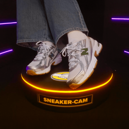 A close-up of a person's feet wearing blue jeans, white socks, and silver and white New Balance sneakers featuring a green "N" logo. They are standing on a small, circular black platform that has a glowing orange ring light around its top edge. The front of the platform displays a yellow label reading "SNEAKER-CAM".