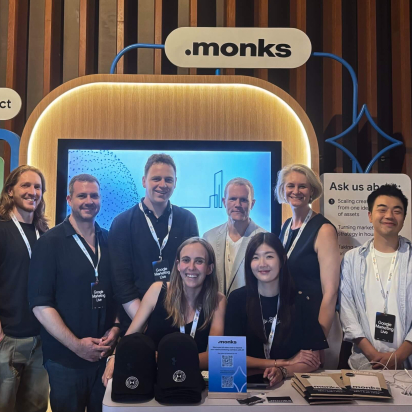 A group of ten smiling professionals posing together at a ".monks" event booth. The attendees are wearing lanyards with "Google Marketing Live" badges. Behind them is a large display screen, an illuminated ".monks" logo, and booth signs reading "Brand impact" and "Ask us about:". In the foreground, a table is set up with branded black caps, brochures, and a small sign featuring a QR code.