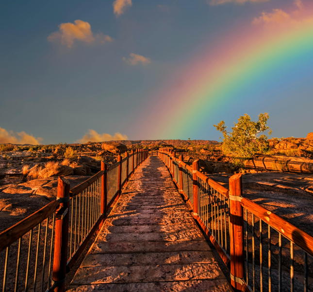 A vibrant rainbow arches over a rustic walkway leading across a rocky landscape bathed in the golden light of sunset. The sky is partly cloudy, enhancing the vividness of the rainbow colors.