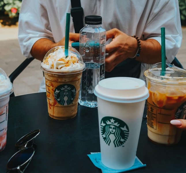 A table outside with all sorts of Starbucks coffe cups on it. At the back of the table you see two hands of a person who is using his mobile.