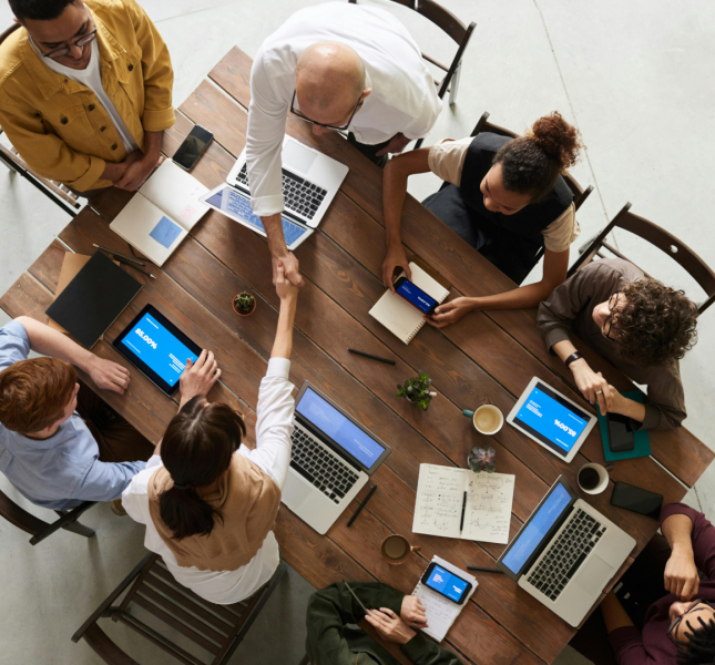 A group of seven people are gathered around a wooden table in a meeting room. They have laptops, tablets, notebooks, and coffee cups in front of them. Two of the individuals are standing and shaking hands over the table, while the others are seated, engaged with their devices or each other. The setting appears collaborative and professional.