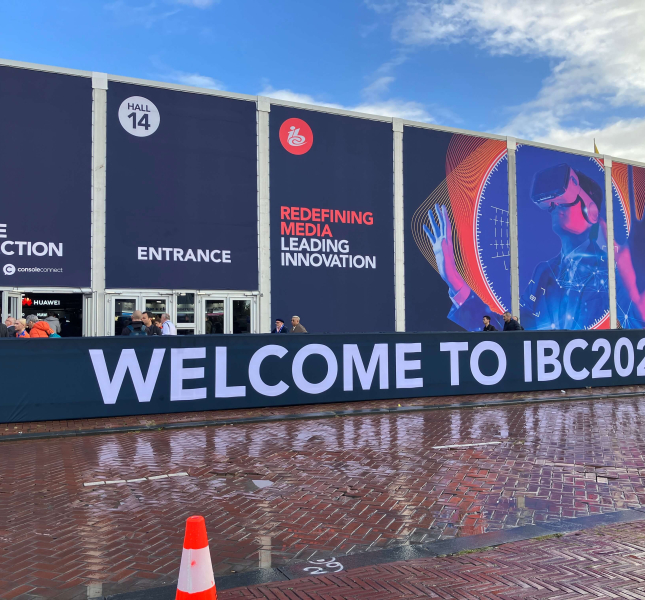The facade of the IBC convention hall is depicted. Large banners outside display text reading "Badge Collection," "Entrance," and "Redefining Media Leading Innovation." A futuristic image of a person wearing a virtual reality headset is featured. The foreground has a sign that reads "Welcome to IBC2024." People are gathered near the entrance, and a traffic cone is visible on the wet pavement.