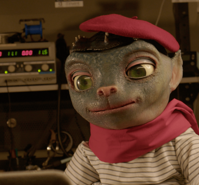Image of Sir Martian, an animatronic in the form of an alien with large eyes and a cat-like nose. He wears an outfit befitting a Parisian street portrait artist.